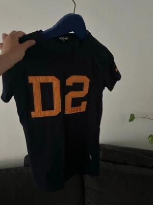 Svart t-shirt med D2-tryck från ICON - Svart t-shirt från ICON med stort orange D2-tryck och liten text 'ICON' på bröstet. Klassisk rund hals och korta ärmar. Perfekt för en avslappnad och sportig stil.