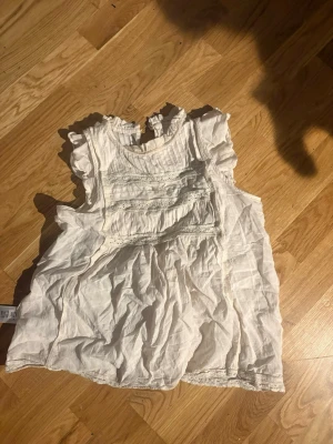 Vit blus med volangdetaljer - Söt vit blus med volangdetaljer vid ärmar och hals. Blusen har spetsinslag och fina veckade partier framtill. Tillverkad i ett lätt och luftigt tyg, perfekt för varmare dagar. Klassisk och romantisk look.