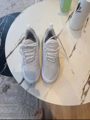 Nike Air Max 270 vita sneakers - Snygga vita Nike Air Max 270 sneakers med mesh-ovandel och tydlig Air Max-dämpning i hälen. Skorna har klassisk snörning, diskret Swoosh-logga på sidan och en modern, sportig look. Perfekta för dig som gillar stilrena och bekväma sneakers.