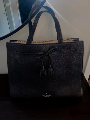 Svart handväska från Kate Spade - Stilren svart handväska från Kate Spade i strukturerat läder. Väskan har två topphandtag, dekorativ knytning med tofsar framtill och guldfärgad dragkedja. Kate Spade-logga i guld längst ner på framsidan. Perfekt för dig som gillar klassisk och elegant design.