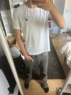 Vit GANT t-shirt med logga - Snygg vit t-shirt från GANT med klassisk logga och sköld på bröstet. T-shirten har rund halsringning och korta ärmar. Tillverkad i mjuk bomull som känns skön mot huden. Perfekt för en clean och enkel stil.