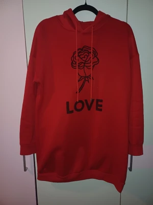 Hoodie - Röd oversized hoodie med huva och dragsnören. Framtill finns ett svart tryck av en ros och texten LOVE. Hoodien har långa ärmar och ribbade muddar. Perfekt för en chill och avslappnad stil.