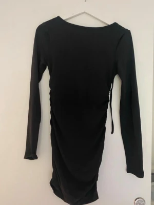 Svart långärmad omlottklänning - Snygg svart klänning med lång ärm och omlottdetalj i midjan. Klänningen har en åtsittande passform och en djup, öppen rygg som ger en trendig look. Perfekt för dig som gillar stilrena och moderna plagg. Aldrig använd 
