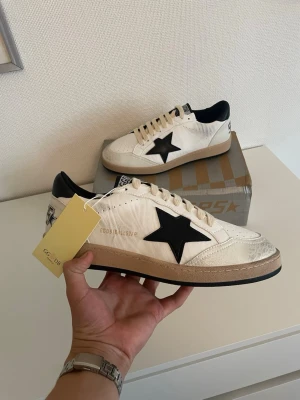 Golden Goose ballstar - Säljer ett par riktigt stilrena Golden Goose i modellen Ball Star. Denna färgkombination har en bas i vitt läder med den ikoniska stjärnan och matchande hälkappa i svart, vilket gör att de passar till i stort sett allt. De har märkets klassiska vintage-look med snygga, fabriksgjorda slitningar. Info om skorna:  Modell: Golden Goose Ball Star  Storlek: 42  Färg: Vit / Svart  Skick: Helt nya och oanvända med lapparna kvar.
