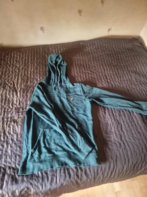 Grön hoodie från Lyle & Scott - Snygg grön hoodie från Lyle & Scott med klassisk gul logga på bröstet. Hoodien har huva med dragsko och en stor magficka framtill. Tillverkad i mjukt bomullsmaterial och har ribbade muddar vid ärmslut och nederkant. Perfekt för en avslappnad stil.