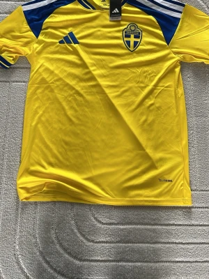 Svensk landslagströja Adidas gul/blå - Gul svensk landslagströja från Adidas med blå detaljer och vita ränder på axlarna. Tröjan har broderat landslagsemblem på bröstet och är tillverkad i lätt funktionsmaterial (Climalite). Perfekt för fotbollsträning eller att bära på matchdag.