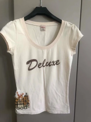 Vit Deluxe t-shirt från Only - Snygg vit t-shirt från Only med texten 'Deluxe' i brunt med prickigt mönster på bröstet. T-shirten har korta ärmar, rund halsringning och en unik detalj med tygpatch och broderi nere vid sidan. Perfekt för en avslappnad och trendig stil. Armhåla till armhåla 38 cm