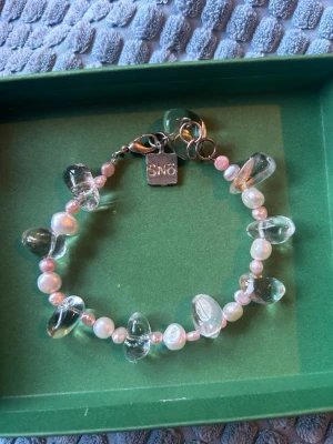 SNO armband med pärlor och stenar - Armband från SNO med små rosa pärlor och större transparenta stenar. Armbandet har ett silverfärgat lås och en liten bricka med SNO-loggan. Perfekt accessoar för dig som gillar en mix av sött och stilrent.