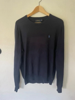 Mörkblå tröja från Polo Ralph Lauren - Klassisk mörkblå tröja från Polo Ralph Lauren med rund halsringning och broderad logga på bröstet. Tröjan har långa ärmar och ribbade muddar vid ärmslut och nederkant. Perfekt för en stilren look.