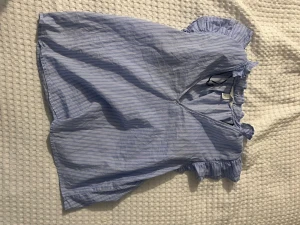 Blårandig blus med volangdetaljer - Söt blårandig blus med vita tunna ränder och volangdetaljer vid ärmarna. Blusen har en v-ringning och är tillverkad i ett lätt bomullstyg som känns luftigt och fräscht. Perfekt för en trendig och avslappnad look.