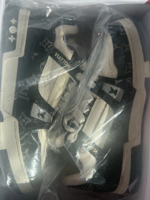 Louis Vuitton svarta och vita sneakers - Snygga Louis Vuitton sneakers i svart och vitt med klassiskt monogrammönster. Skorna har vita detaljer, grov sula och LV-logga på sidan. Tillverkade i en mix av läder och textil för en exklusiv känsla. Perfekta för dig som vill sticka ut med lyxig streetstyle.