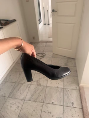 Svarta klassiska pumps med blockklack - Stilrena svarta pumps med rund tå och stabil blockklack. Skorna har en enkel och tidlös design som passar till många olika outfits. Tillverkade i slätt material med diskreta sömmar längs kanten. Perfekta för dig som gillar en klassisk look.