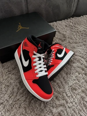 Air Jordan 1 Mid röd svart vit - Säljer ett par Air Jordan 1 Mid sneakers i rött, svart och vitt. Skorna har klassisk hög siluett, vit swoosh och röda detaljer i läder. Tungan är svart med röd Jumpman-logga och vita skosnören. Perfekta för dig som gillar streetwear och ikonisk design.