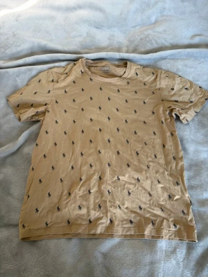 Beige t-shirt från Polo Ralph Lauren - Säljer en beige t-shirt från Polo Ralph Lauren med små mörkblå loggor tryckta över hela tröjan. Klassisk rund halsringning och korta ärmar. Tillverkad i mjuk bomull som känns skön mot huden. Perfekt för en avslappnad och stilren look.