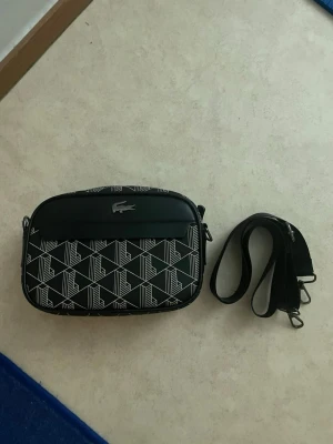 Lacoste bag  - Snygg svart axelväska från Lacoste med vitt geometriskt mönster och klassisk krokodillogga framtill. Väskan har avtagbar axelrem och dragkedja upptill. Tillverkad i slitstarkt syntetmaterial, perfekt för att bära dina viktigaste grejer.