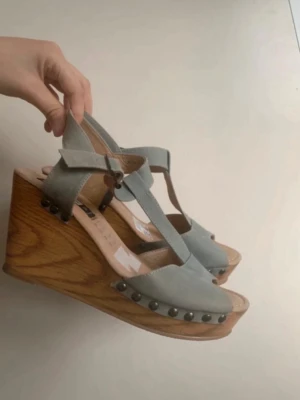 Gråa kilklackade sandaler - Snygga sandaler med grått läder och kilklack i trälook. Dekorerade med svarta nitar längs sulan och har T-rem med spänne vid ankeln. Perfekta för dig som vill ha både höjd och stil i sommarens outfit. Helt nya. 