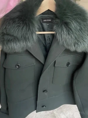 Mörkgrön pälsjacka från Meotine - Stilren mörkgrön pälsjacka från Meotine med stor fluffig fuskpälskrage. Jackan har två stora bröstfickor med lock och knappar framtill. Kortare modell med breda ärmar och markerad axel. Perfekt för kalla vinterdagar och ger en lyxig känsla till din outfit.