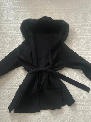 Svart kappa med fuskpäls och huva - Stilren svart kappa med stor huva kantad av mjuk fuskpäls. Kappan har knytskärp i midjan och två stora fickor framtill. Perfekt för kyliga dagar när du vill vara både varm och snygg. Materialet känns mjukt och tjockt, vilket ger en lyxig känsla.