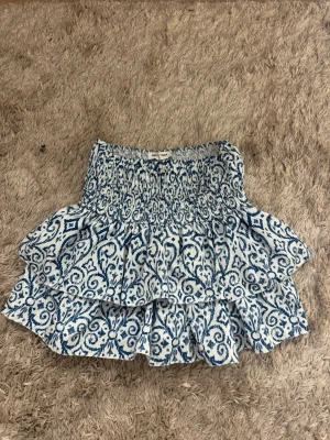 Blåvit volangkjol  - Supersöt kort kjol från MEET x TIGER i blått och vitt med volanger och smock i midjan. Kjolen har ett unikt mönster och är perfekt för dig som gillar en lekfull och trendig stil. Tillverkad i ett lätt och luftigt material som passar till många outfits.