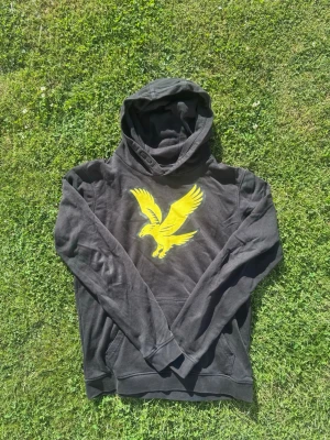 Svart hoodie med gul örntryck - Säljer en svart hoodie med stor gul örn på bröstet. Hoodien har huva, känguruficka framtill och ribbade muddar vid ärmar och nederkant. Perfekt för dig som gillar streetwear och vill sticka ut med ett coolt tryck.