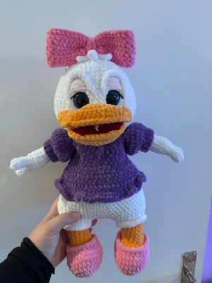 Virkad Daisy Duck gosedjur - Supergullig handvirkad Daisy Duck i mjukt garn. Hon har en stor rosa rosett, lila tröja och rosa skor. Perfekt som present eller dekoration för Disney-fans. Färgerna är vit, lila, rosa och orange. Gosedjuret är detaljerat och har stora glittriga säkerhetsögon.