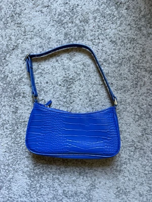 Blå axelväska i krokopräglat material - Snygg blå handväska med axelrem och krokopräglat mönster. Väskan har en stilren, avlång form och stängs med dragkedja. Perfekt för dig som vill ha en trendig accessoar som sticker ut.
