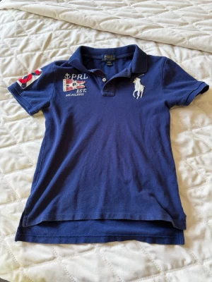 Marinblå pikétröja från Polo Ralph Lauren - Snygg marinblå pikétröja från Polo Ralph Lauren med klassisk krage, broderad logga på bröstet och detaljerade patches på bröst och ärm. Tröjan är i bomull och har korta ärmar samt knappar vid halsen. Perfekt för en sportig och stilren look.