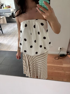 Vit prickig tubtopp i satin - Säljer en vit axelbandslös tubtopp i glansigt satinmaterial med stora svarta prickar. Toppen har en bandeauform och sitter löst över kroppen för en chill och trendig look. Perfekt att styla med kjol eller jeans.