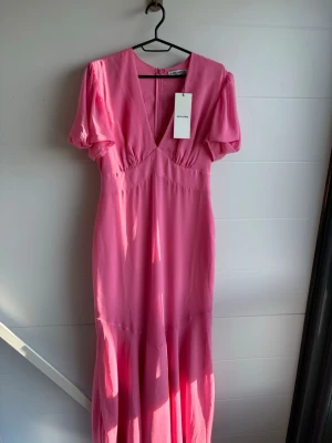 Hot pink maxiklänning från Adoore - Säljer en snygg hot pink maxiklänning från Adoore med v-ringning och korta puffärmar. Klänningen har en figurnära passform upptill och faller snyggt nedtill med ett lätt, flowy tyg. Perfekt för dig som vill sticka ut med färg och stil.