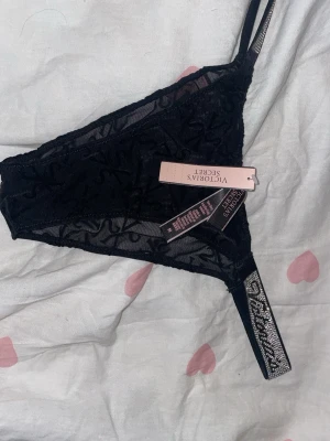 Svarta spets-trosor Victoria's Secret - Säljer ett par svarta trosor från Victoria's Secret i transparent mesh med broderat VS-mönster. Sidorna har en glittrig detalj med text och trosorna har en feminin, låg skärning. Perfekta för dig som gillar snygga och trendiga underkläder.