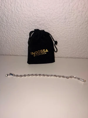 Flätat silverarmband från Indossa - Snyggt flätat armband i silverfärg från Indossa by Sweden. Armbandet har en elegant tvinnad design och kommer med en svart tygpåse med guldfärgad logga. Perfekt accessoar för att lyfta din stil.