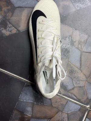 Nike Mercurial vita fotbollsskor - Säljer ett par Nike Mercurial fotbollsskor i vitt med svart swoosh och färgglada detaljer i rött, rosa och mintgrönt på hälen. Skorna har snörning och ovandel i textilmaterial för bra passform och grepp på planen. Perfekta för dig som vill sticka ut på fotbollsplanen.