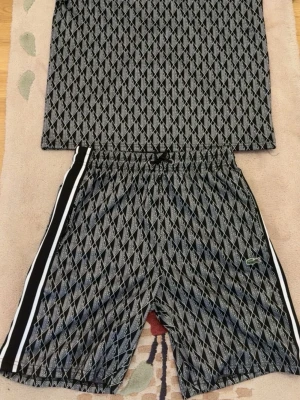 Svarta mönstrade shorts från Lacoste - Säljer ett par svarta shorts från Lacoste med vitt geometriskt mönster och vita ränder längs sidorna. Shortsen har snörning i midjan och är perfekta för en sportig look. Klassisk Lacoste-logga på benet och bekväm passform. Helt ny aldrig använd