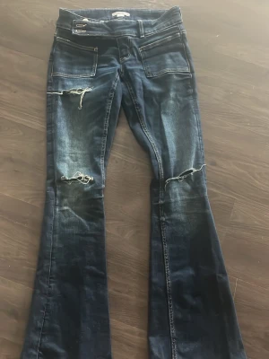 Mörkblå bootcut jeans med slitningar - Säljer ett par mörkblå bootcut jeans med slitna detaljer och hål vid knäna. Jeansen har framfickor, dragkedja och knapp i midjan samt ljusa tvättade partier. Perfekta för en avslappnad och trendig look.