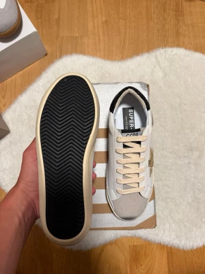 Vita sneakers från Superga - Snygga vita sneakers från Superga med beige detaljer och svart hälflik. Skorna har en klassisk låg siluett, beige snörning och en svart sula med fiskbensmönster undertill. Materialet är en mix av textil och mocka för en trendig look.
