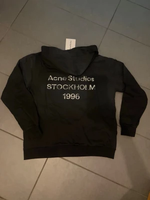 Svart hoodie Acne Studios Stockholm M - Säljer en svart hoodie från Acne Studios med texten 'Acne Studios STOCKHOLM 1996' i vitt på ryggen. Klassisk passform med huva och ribbade muddar. Perfekt för dig som gillar stilrena och trendiga plagg från populära märken.