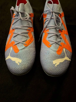 Puma blå-orange fotbollsskor Bra skick storlek 41  - Snygga fotbollsskor från Puma i ljusblått med orange detaljer och vita Puma-loggor. Skorna har en stickad ovandel för bra passform och snörning för stabilitet. Perfekta för dig som vill sticka ut på planen med både 