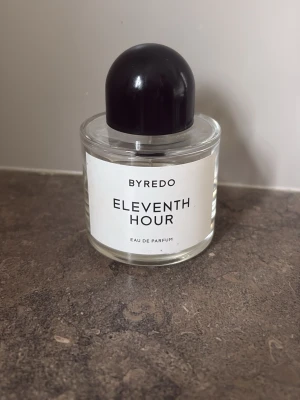 Byredo Eleventh Hour Eau de Parfum - Byredo Eleventh Hour Eau de Parfum i en stilren, rund glasflaska med svart rund kork. Flaskan har en vit etikett med svart text och minimalistisk design. Perfekt för dig som gillar exklusiva och moderna dofter. 100 ml flaska och ca 45-50 ml kvar.