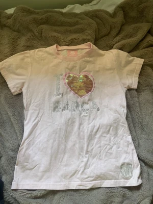 Rosa FC Barcelona t-shirt  - Söt ljusrosa t-shirt från FC Barcelona med korta ärmar och ett stort paljetthjärta på bröstet. Texten 'I ❤️ BARÇA' i glittriga stenar och klubbens logga nere vid fållen. Tappat två stenar men annars i bra skick. Står storlek L men passar som S