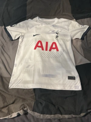 Tottenham Hotspur matchtröja Nike - Vit Tottenham Hotspur matchtröja från Nike med röda AIA-loggan på bröstet, klubbmärke och Nike-logga i mörkblått. Tröjan har subtila grafiska mönster, mörkblå detaljer vid ärmslut och sponsorlogga på ärmen. Perfekt för fotbollsfans!
