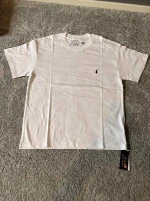 Ralph lauren T-shirt Vit Ny-oanvänd (M) -  Säljer en helt ny och oanvänd Ralph Lauren T-shirt i storlek M. Stilren och tidlös T-shirt i vit färg med den klassiska svarta Ralph Lauren-loggan broderad på bröstet. Tillverkad i mjukt och bekvämt material av hög kvalitet – perfekt för både vardag och en avslappnad, stilren look. 🔹 Märke: Ralph Lauren 🔹 Modell: T-shirt 🔹 Färg: Mörkblå med vit logga 🔹 Storlek: M 🔹 Skick: Helt ny / oanvänd 🔹 Passform: Klassisk Nypris är högt – perfekt möjlighet till ett fynd