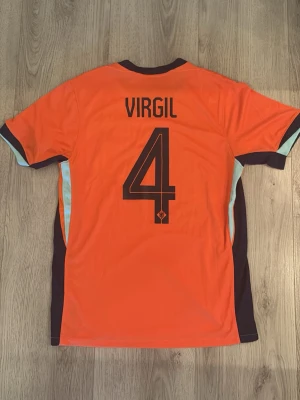 Holland fotbollströja Nike Virgil #4 - Snygg orange Holland fotbollströja från Nike med svarta detaljer och nummer 4 samt namnet Virgil på ryggen. Tröjan har korta ärmar, svart Nike-logga och landslagets emblem på bröstet. Perfekt för dig som vill stötta Nederländerna på match!
