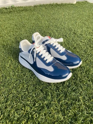 Prada skor  - Snygga sneakers i blått och vitt med glansiga lackdetaljer och meshpaneler för extra andningsförmåga. Skorna har vita snören, vit sula och en röd detalj på plösen. Perfekta för dig som gillar en sportig och fräsch stil.