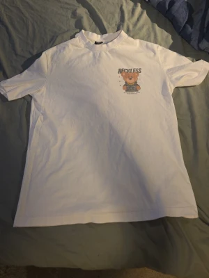 Vit t-shirt med fet tryck  - Säljer en vit t-shirt med ett litet tryck på bröstet där det står 'RECKLESS' och en tecknad björn. T-shirten har relaxed fit, rund hals och är kortärmad. Perfekt för dig som gillar streetwear och vill ha något chill i garderoben.