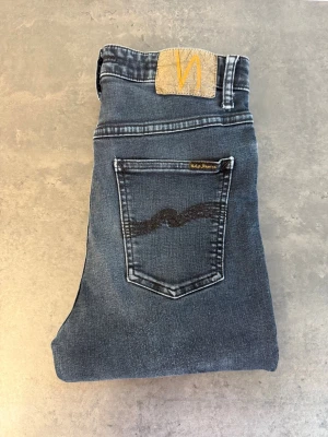 Grå slim jeans från Nudie Jeans W30 L32 - Snygga grå slimjeans från Nudie Jeans med klassisk femficksdesign och gul logotyppatch bak. Jeansen har en mörk tvätt och den ikoniska vågdetaljen på bakfickan. Perfekta för en avslappnad och trendig look.