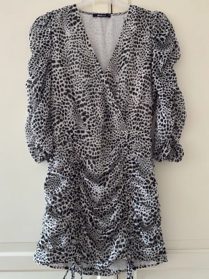 Leopardmönstrad klänning från Gina Tricot - Kort svartvit leopardmönstrad klänning från Gina Tricot med v-ringning, puffiga långärmar och omlottdesign. Klänningen har rynkade detaljer och snörning nertill för en snygg siluett. Med dragkedja på sidan. Storlek 38. I fint skick. 
