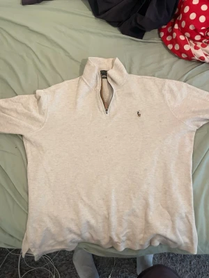  quarter zip från Polo Ralph Lauren - Säljer en ljusgrå långärmad quarter zip från Polo Ralph Lauren med klassisk broderad logga på bröstet. Tröjan har hög krage med dragkedja och är tillverkad i mjukt bomullsmaterial. Perfekt för en avslappnad och stilren look.
