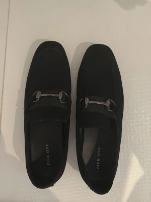 Svarta loafers från Pier One - Stilrena svarta loafers från Pier One med dekorativt metallspänne framtill. Skorna har en klassisk form och är tillverkade i mockaliknande material med gummisula. Perfekta för dig som gillar en clean och tidlös look. Använda endast 2 gånger. 