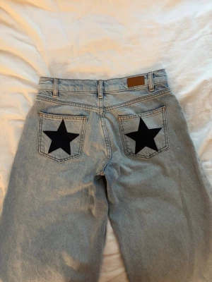 Ljusblå jeans med svarta stjärnor - Säljer ett par ljusblå jeans från subdued med raka ben och coola svarta stjärnor på bakfickorna. Klassisk femficksmodell med hög midja och bälteshällor. Jeansen är i ett mjukt denimtyg som känns skönt att bära.