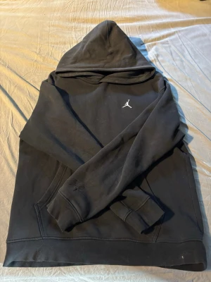 Svart Jordan hoodie med logga - Säljer en svart hoodie från Jordan med den klassiska Jumpman-loggan broderad på bröstet och Michael Jordans signatur på ärmen. Hoodien har huva och magficka, samt ribbade muddar vid ärmslut och nederkant. Perfekt för en sportig och avslappnad stil.
